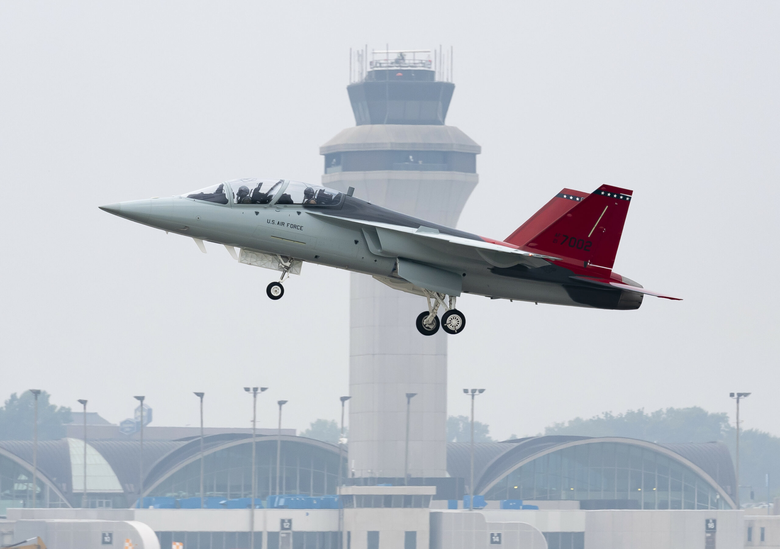 A T-7A Red Hawk is seen taking off from an airport. The words "US Air Force" are painted on its body, on the side of the cockpit. Its red tails have the number 7002 painted in black. An airport tower is seen in the background.