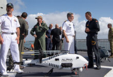 Thirteen uniformed personnel from different units converse during a NATO maritime exercise. A white unmanned helicopter drone is on the foreground, with the words "Alpha Unmanned Systems" painted on its body and "A900" on its tail. The sea and sky is visible on the background.