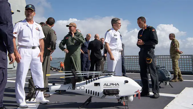 Thirteen uniformed personnel from different units converse during a NATO maritime exercise. A white unmanned helicopter drone is on the foreground, with the words "Alpha Unmanned Systems" painted on its body and "A900" on its tail. The sea and sky is visible on the background.