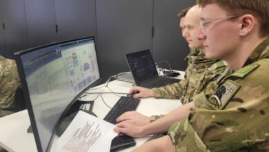 Three uniformed personnel from the New Zealand Defence Force huddle in front of a computer screen displaying programs most likely used for cyber defense purposes. An officer, with their back turned, is on the background. A keyboard, mouse, and laptop are on the computer desk, alongside a piece of paper with a bar graph printed on it.