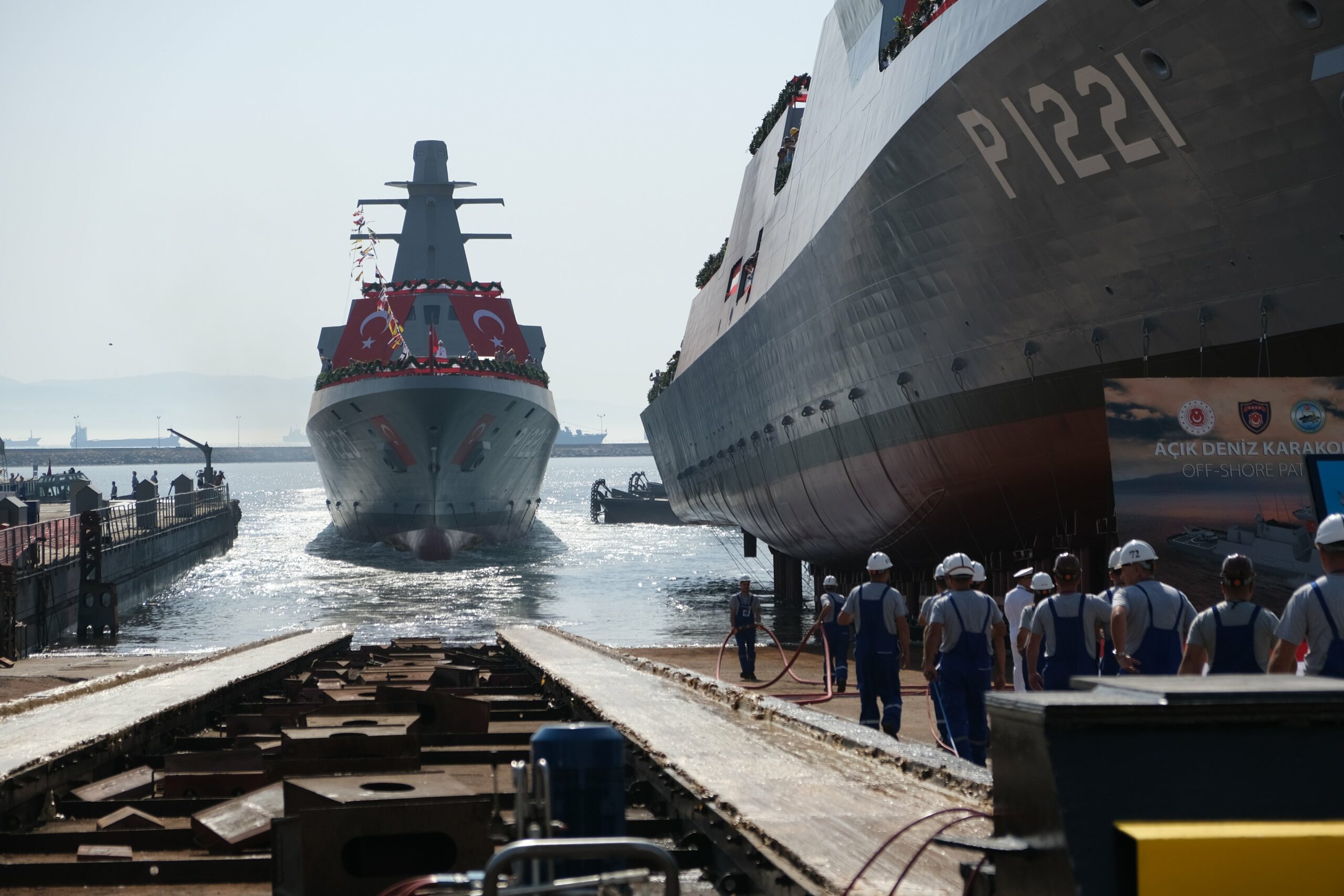 Turkey's new offshore patrol vessels, AKHİSAR ( P-1220) and KOÇHİSAR (P-1221) are seen on a dock, with P-1220 already on the water and P-1221 on the ready for launch. Dock workers can be seen on the ground, by the side of P-1221. Both ships are adorned with wreaths and flags of Turkey.