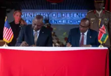 Secretary of Defense Lloyd J. Austin III (left) and Kenyan Defense Minister Aden Bare Duale (right) sit beside each other while signing the papers of the five-year framework for defense cooperation. They are flanked by one military personnel each on the background. A US flag is placed on Austin's side, while a Kenyan flag is on Duale's.