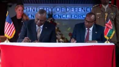 Secretary of Defense Lloyd J. Austin III (left) and Kenyan Defense Minister Aden Bare Duale (right) sit beside each other while signing the papers of the five-year framework for defense cooperation. They are flanked by one military personnel each on the background. A US flag is placed on Austin's side, while a Kenyan flag is on Duale's.