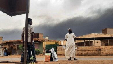 Smoke rises above buildings as people wait on the side of a road with some belongings, in Khartoum