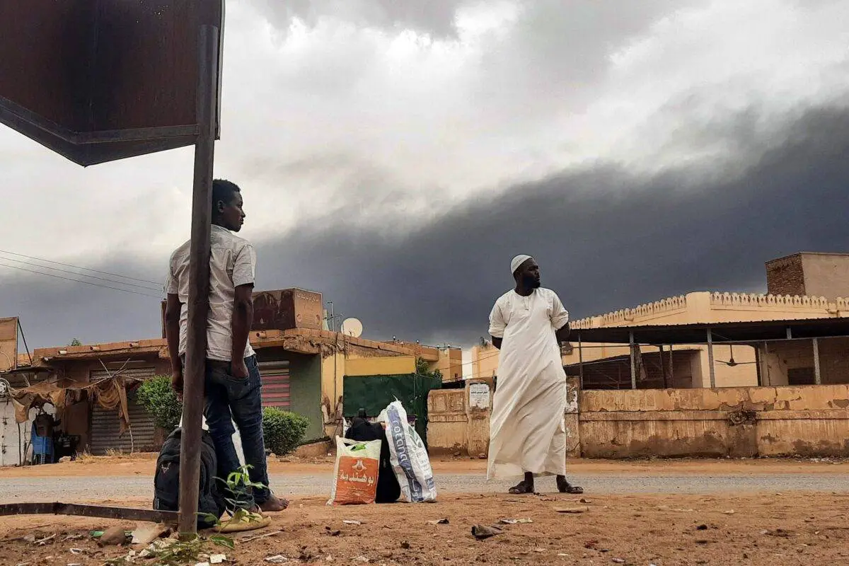Smoke rises above buildings as people wait on the side of a road with some belongings, in Khartoum