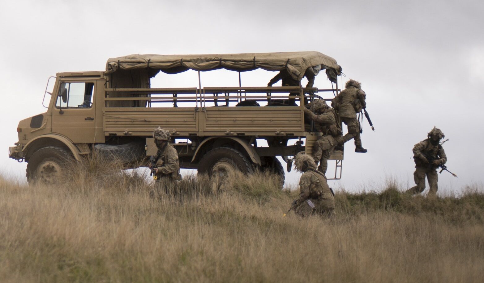A stationary Mercedes Benz Unimog is seen in a grassland. Around its back, three camouflaged soldiers surround it and three more are jumping out of it.