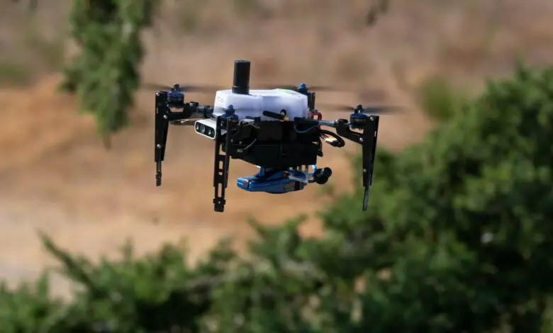 A small hovering drone is front and center in the picture. In the blurred out background, some greenery are seen in an otherwise rocky/sandy area.