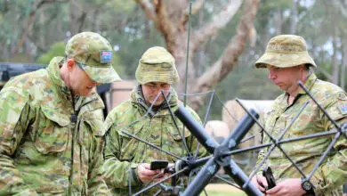 Three soldiers in camo uniform test out a hihg-frequency device. They are huddled together in the middle of the frame.