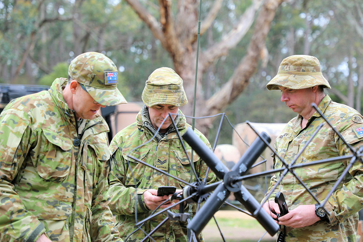 Three soldiers in camo uniform test out a hihg-frequency device. They are huddled together in the middle of the frame.