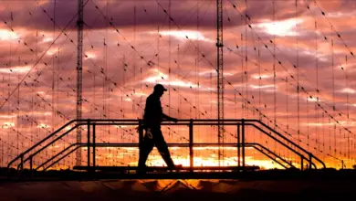 A man crosses a bridge in a Jindalee Operational Radar Network (JORN) site in Australia. In the background, two cable towers are connected to hundreds of cable lines, suggesting that all of it is part of the radar network itself. Everything is seen in the image as shadowy figures as a red-orange sunset is seen casting its light from the far background. Overcast clouds cover the whole sky.