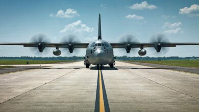 A Lockheed Martin C-130J-30 Super Hercules plane is seen front and center on the image. Its four wing propellers are spinning as it appears to be stationary on an airplane runway.