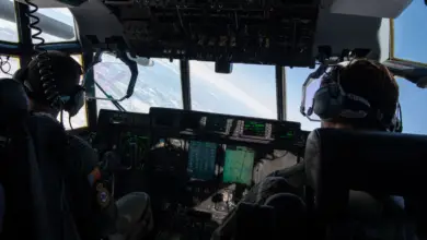 US Air Force pilots are flying an aircraft above the clouds. The photo was taken inside the cockpit, between the two uniformed personnel.