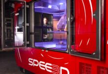 3D printing company SPEE3D's printing equipment, WarpSPEE3D, is seen on the photo. A man looks on in the background as he operates it, showing that the machine is the size of a small room, approximately 3 meters in height and 4 meters in length. Huge panels of glass between its sides show how it prints metal inside.