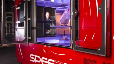 3D printing company SPEE3D's printing equipment, WarpSPEE3D, is seen on the photo. A man looks on in the background as he operates it, showing that the machine is the size of a small room, approximately 3 meters in height and 4 meters in length. Huge panels of glass between its sides show how it prints metal inside.
