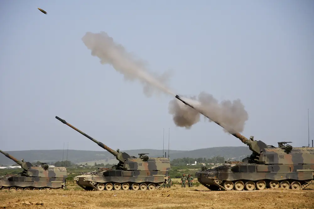 Three tanks, facing left, all have its main gun pointed upwards at a 45-degree angle. The rightmost tank, which is also closest, is in the process of firing ammunition. Smoke is seen shooting out of the gun, with the blurred ammunition in front.