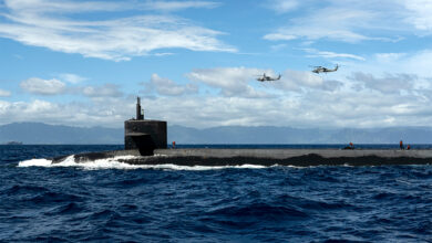 USS Henry M. Jackson, a US submarine, is seen breaking the surface in the middle of the image. In the background, two helicopters are flying overhead. The sky is blue and cloudy.