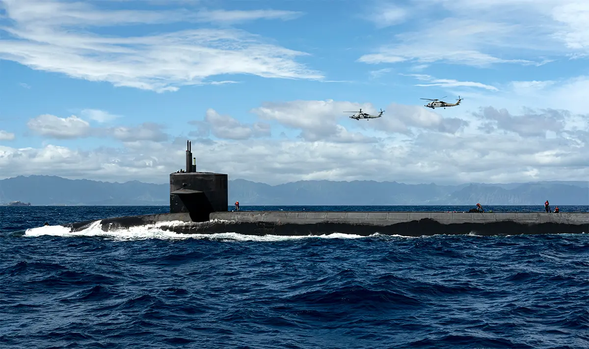 USS Henry M. Jackson, a US submarine, is seen breaking the surface in the middle of the image. In the background, two helicopters are flying overhead. The sky is blue and cloudy.