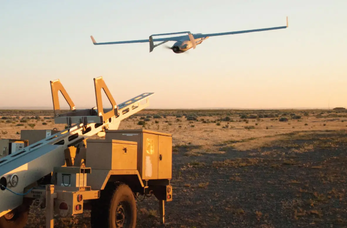 An Insitu Integrator drone is seen taking off, seemingly launched by a wheeled launcher on the left side of the image. The sunset tinges the whole scenery with a bright, warm orange hue.
