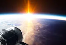 A satellite is seen in space, on the lower right corner of the image, The earth is seen on the background, and the sun is rising on its horizon.