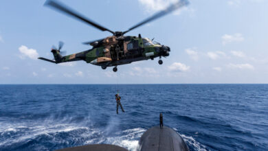 A Royal Australian Navy sailor is lowered to the casing of HMAS Collins from an MRH-90 Maritime Support Helicopter while sailing in the Bay of Bengal. The background is a clear blue sky with a few white clouds. The deep blue water below is distrubed by the gusts from the helicopter blades.