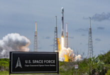 A rocket launches from at Cape Canaveral Space Force Station, Florida. On the left of the foreground, a sign that says "U.S. SPAC FORCE Cape Canaveral Space Force Station" is seen. The Space Force Logo is on the left of the sign.