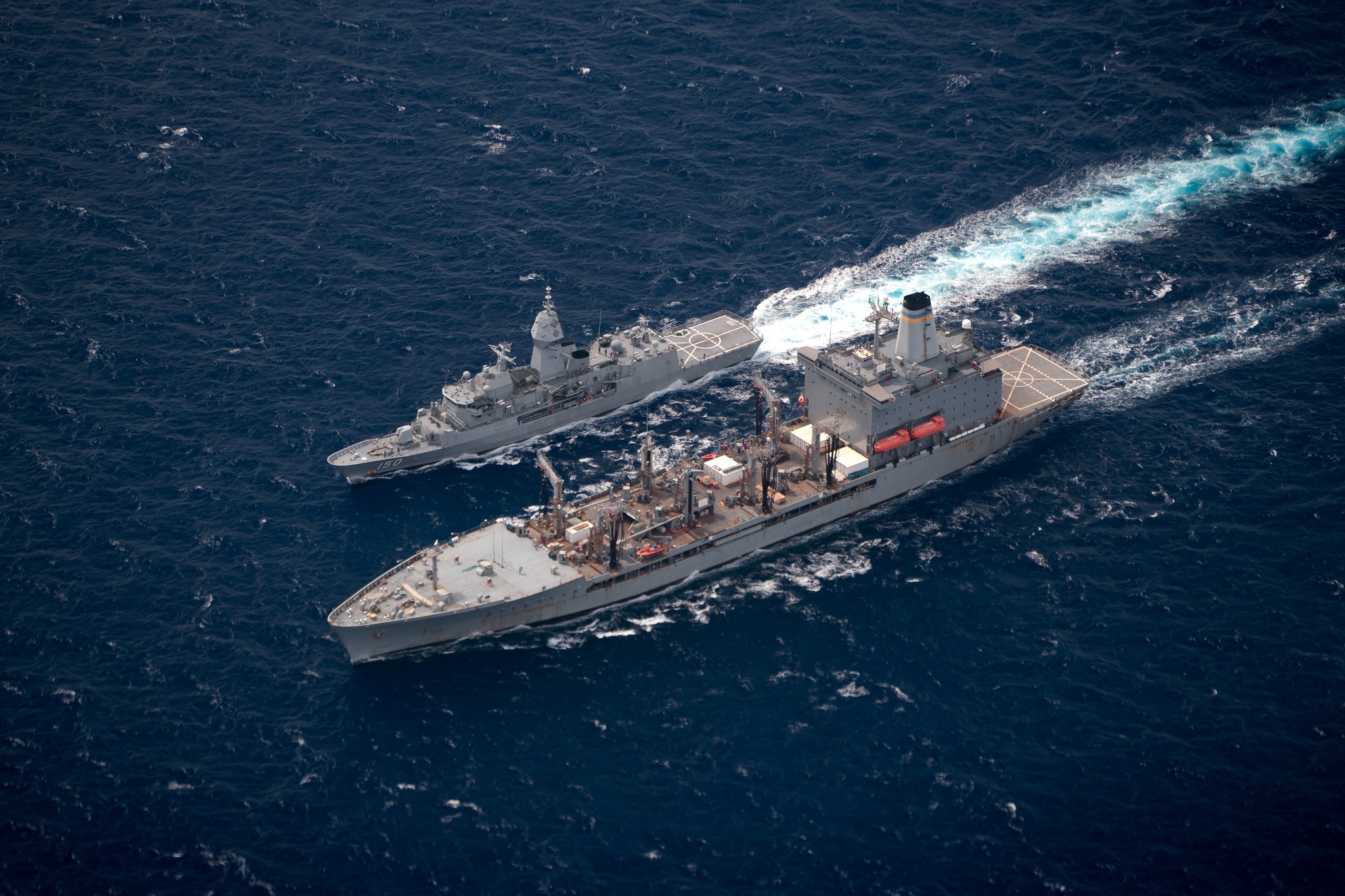 HMAS Anzac III sails alongside USNS Rappahannock. The photo is taken at a bird's eyeview. The sea is a deep blue.