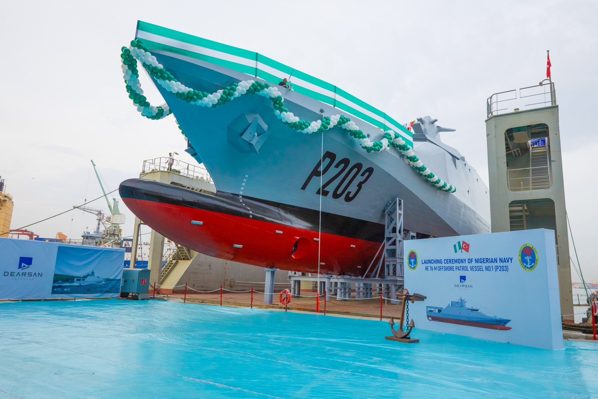 The new Nigerian Navy offshore patrol vessel, P203, is seen in a shipyard in Istanbul, Turkey. A rope of balloons decorate its deck's perimeter and a sign that says "LAUNCHING CEREMONY OF NIGERIAN ARMY HE 76M OFFSHORE PATROL VESSEL NO. 1 (P203)" is seen erected on the ground. The ship's grey hull has the name P203 painted in black, on the left.