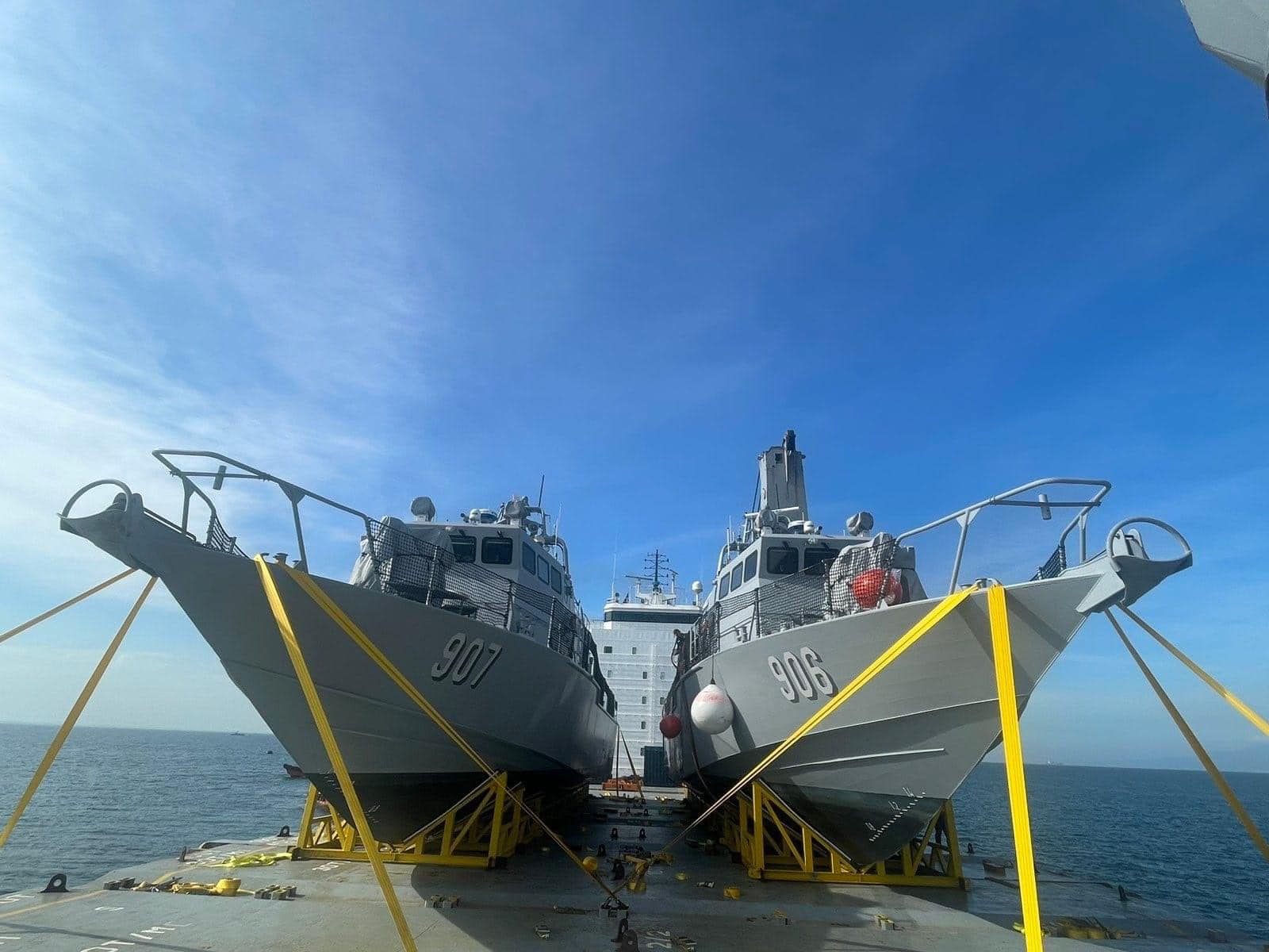 Fast-attack interdiction crafts (FAIC) PG-906 and PG-907 are seen docked in a port, both with two yellow ropes from the ground each tied over their hulls. Their names are painted in white on the side of their bodies. The blue sky acts as a background, with the port's blue-green water peeking on the lower back of the image.
