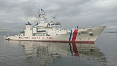 BRP Teresa Magbanua (MRRV-9701) is seen on calm, grey waters. The sky is overcast. The white ship has '9701' painted on its right side in black. The words 'PHILIPPINE COAST GUARD' is written across its body in red. A thin blue line and a thicker red line is painted close to the ship's front.