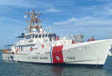 The US Coast Guard's Sentinel-class cutter, USCGC Melvin Bell (WPC-1155), is seen sailing the water. The ship is painted white, with the words "U.S. COAST GUARD" painted on the side of the hull in black. Close to the front, "1155" is also painted. Separating the two are a thin stripe of blue and a thick stripe of red, where the coast guard's coat of arms is located. Nine crew members are seen on deck. A fair, sparsely-cloudy blue sky serves as the image's background. The water is a deep blue-green color.