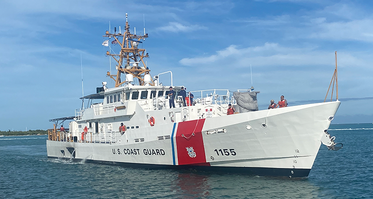 The US Coast Guard's Sentinel-class cutter, USCGC Melvin Bell (WPC-1155), is seen sailing the water. The ship is painted white, with the words "U.S. COAST GUARD" painted on the side of the hull in black. Close to the front, "1155" is also painted. Separating the two are a thin stripe of blue and a thick stripe of red, where the coast guard's coat of arms is located. Nine crew members are seen on deck. A fair, sparsely-cloudy blue sky serves as the image's background. The water is a deep blue-green color.