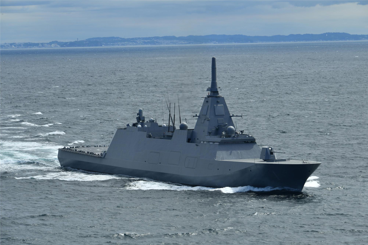 JS Mogami (FFM-1) is seen sailing in open water. A small strip of land serves as the distant background of the image. The ship's hull is colored grey, and the water is a dull, grayish blue.
