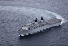 HMS Albion, an amphibious warfare ship, is seen sailing blue-grey waters. The photo appears to have been taken from above the waters, possible from an aircraft. The ship is painted grey and has "L14" painted on its side in black.