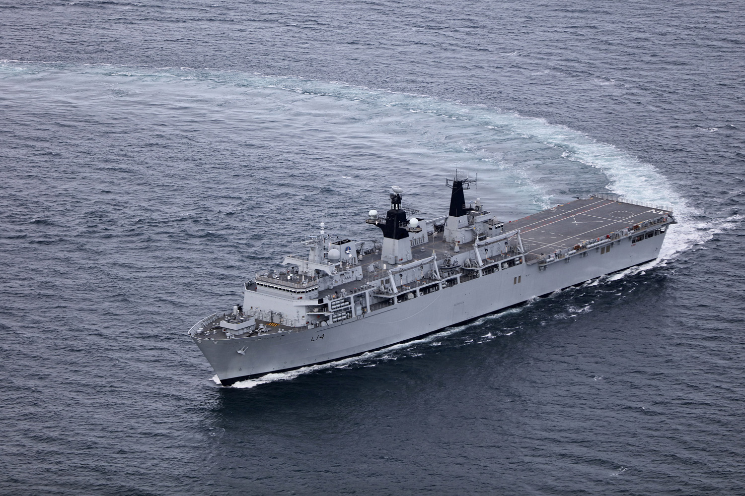 HMS Albion, an amphibious warfare ship, is seen sailing blue-grey waters. The photo appears to have been taken from above the waters, possible from an aircraft. The ship is painted grey and has "L14" painted on its side in black.