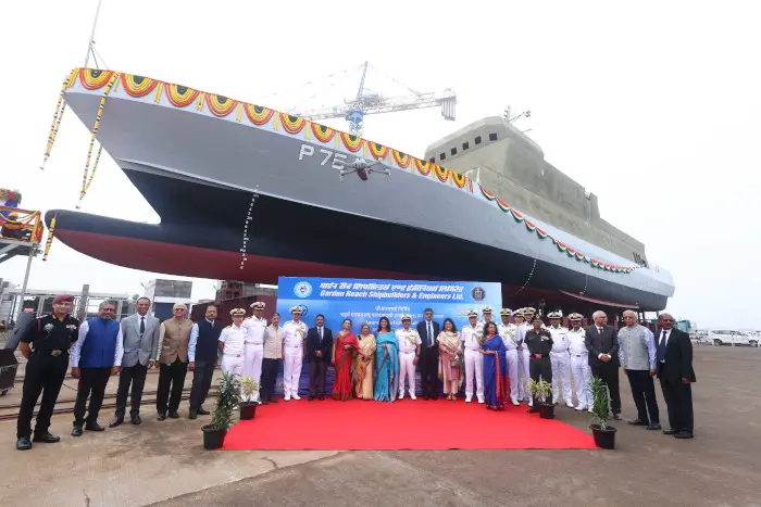 ASW-SWC Amini (P75), an Arnala-class anti-submarine corvette, is seen docked in the background. The photo was taken during its launching ceremony, and a group of 28 people are lined up in the foreground. They appear to be officials from the Indian Navy and the shipyard the corvette was built in. A red carpet is laid out in the middle and a banner for the event is displayed behind them.
