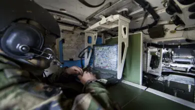A military personnel, wearing camouflage and a microphoned headset, is seen using a laptop displaying a map. The photo appears to be taken inside a cramped military vehicle.