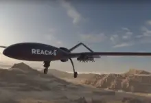 A 3D rendering of EDGE's REACH-S unmanned aerial vehicle (UAV) is seen flying over a dry, mountainous desert landscape. The sky is a dull blue with the sun casting light shadows as it begins to set offscreen.