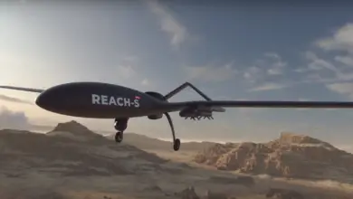 A 3D rendering of EDGE's REACH-S unmanned aerial vehicle (UAV) is seen flying over a dry, mountainous desert landscape. The sky is a dull blue with the sun casting light shadows as it begins to set offscreen.