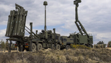 Sky Sabre, a state-of-the-art air defence system, is seen stationed in a grassy plain. Two trucks face each other, carrying various radar/sensor-looking equipment on their backs. On the far left and right side of the background, a tank is seen on each side. The sky is cloudy and overcast.