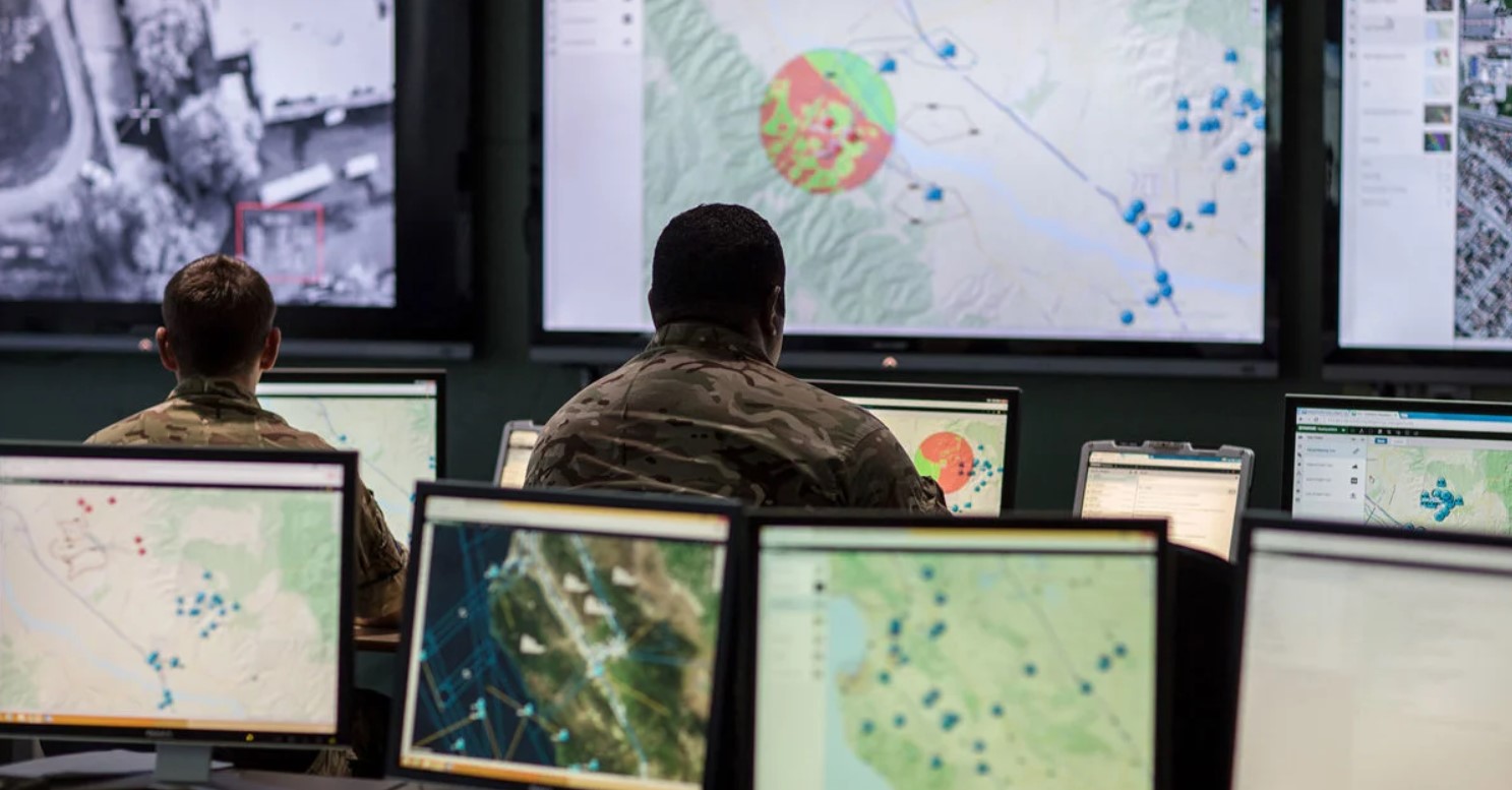 Two military personnel wearing camouflage uniform look at their computer screens displaying what appears to be maps. They're facing the background, which has three large screens displaying different maps. Similarly, four smaller screens make up the foreground.