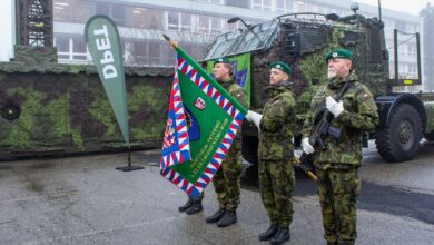 Four soldiers in camouflage stand in attention, facing left. The third from the left is holding a green flag that appears to be the standard banner of an army regiment. It's bordered by a frame with red, white and blue triangle patterns. The fourth soldier is holding a rifle close to his torso. BEhind them, a military truck is parked.