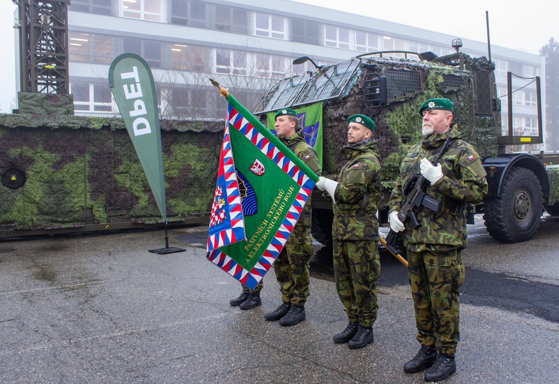 Four soldiers in camouflage stand in attention, facing left. The third from the left is holding a green flag that appears to be the standard banner of an army regiment. It's bordered by a frame with red, white and blue triangle patterns. The fourth soldier is holding a rifle close to his torso. BEhind them, a military truck is parked.