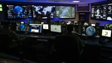 The dimly-lit room of the US Space Command Joint Operations Center is seen in the photo. The far left wall has three large monitors mounted on it, while the far right wall has one visible. The room is full of desks with uniformed personnel working on their computers. Most of the screens, including the walls' TVs', are displaying various interfaces that include maps and graphs.