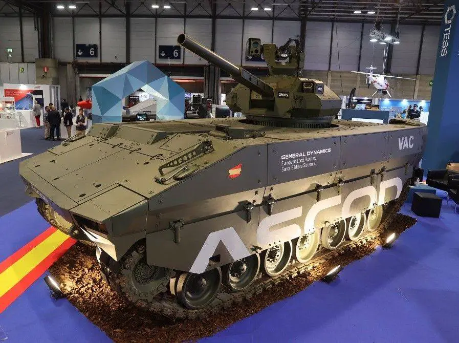 The Spanish Army's Vehículo de Apoyo de Cadenas (VAC) is seen on display in what appears to be a weapons expo, inside a large building with blue carpet. The tank is colored deep green.