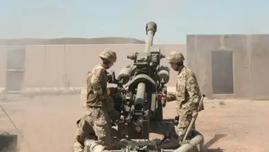 Three soldiers tend to a large-caliber weapon system that has its barrel pointed over towards the horizon. They are wearing camouflage uniforms and their faces are blurred. The landscape is dry land, and the sun is shining bright out of frame. Separating the soldiers and the background is a red-brown wall with a concrete shelter on the right side.