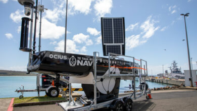 The Bluebottle is seen on a mobile wheeled platform, seemingly beign prepared for delivery. Its sail is propped up, with solar panels visibly attached to it. The black body of the vessel has "OCIUS", "Te Taua Moana Navy" (with their logo beside it), and "BLUEBOTTLE" painted on it in white. The background appears to be a port, with a grey ship on the far right side.