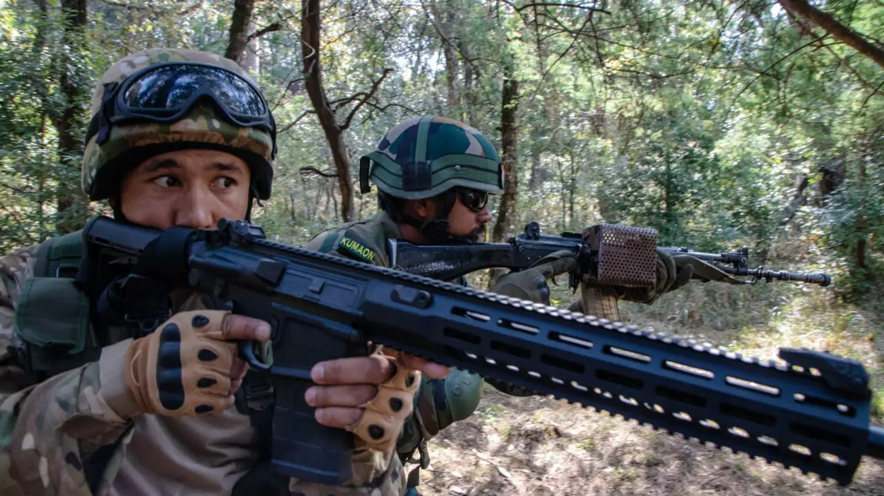 Two soldiers are seen patrolling in what appears to be a forest in daylight with their rifles at the ready. Both of them are pointing their weapons to the right. The one on the foreground, positioned on the left, is holding a Sig Sauer rifle. The other soldier is positioned behind his left, on the center of the image. They are both wearing camouflage.