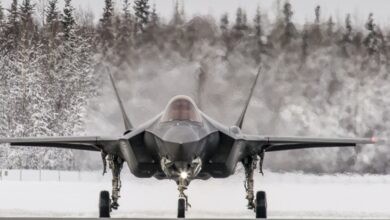 A black F-35 fighter jet plane is seen grounded, facing the viewer. The area it is in is snowy, with the ground completely covered in white snow. The background is a forest of snow-covered trees. The background on the back of the plane is blurred, presumably due to the heat distortion emitted by the plane.