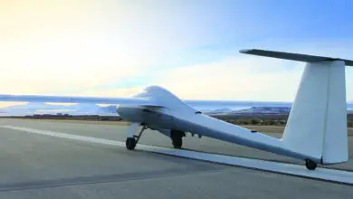 An ULTRA Long Endurance Uncrewed Aerial System is seen grounded on what appears to be a military runway. The drone is shaped like a sleeker aircraft, with a thin body, tail, and pair of wings. It's painted a light grey color. The background is a clear blue sky, with a lot of orange light on the left side of the image, probably caused by the rising of the sun.