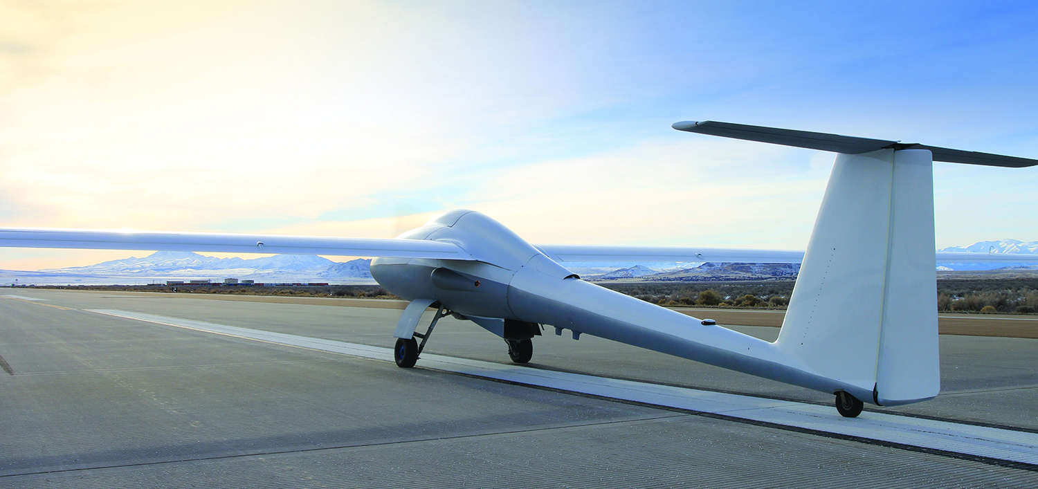 An ULTRA Long Endurance Uncrewed Aerial System is seen grounded on what appears to be a military runway. The drone is shaped like a sleeker aircraft, with a thin body, tail, and pair of wings. It's painted a light grey color. The background is a clear blue sky, with a lot of orange light on the left side of the image, probably caused by the rising of the sun.